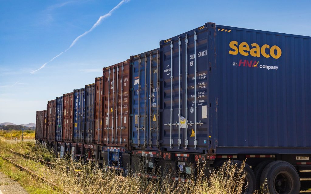 Row of vibrant cargo containers on train tracks under a clear blue sky.